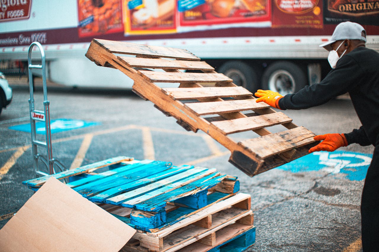 Worker with mask stacking pallets in an outdoor setting. Focus on safety and labor.